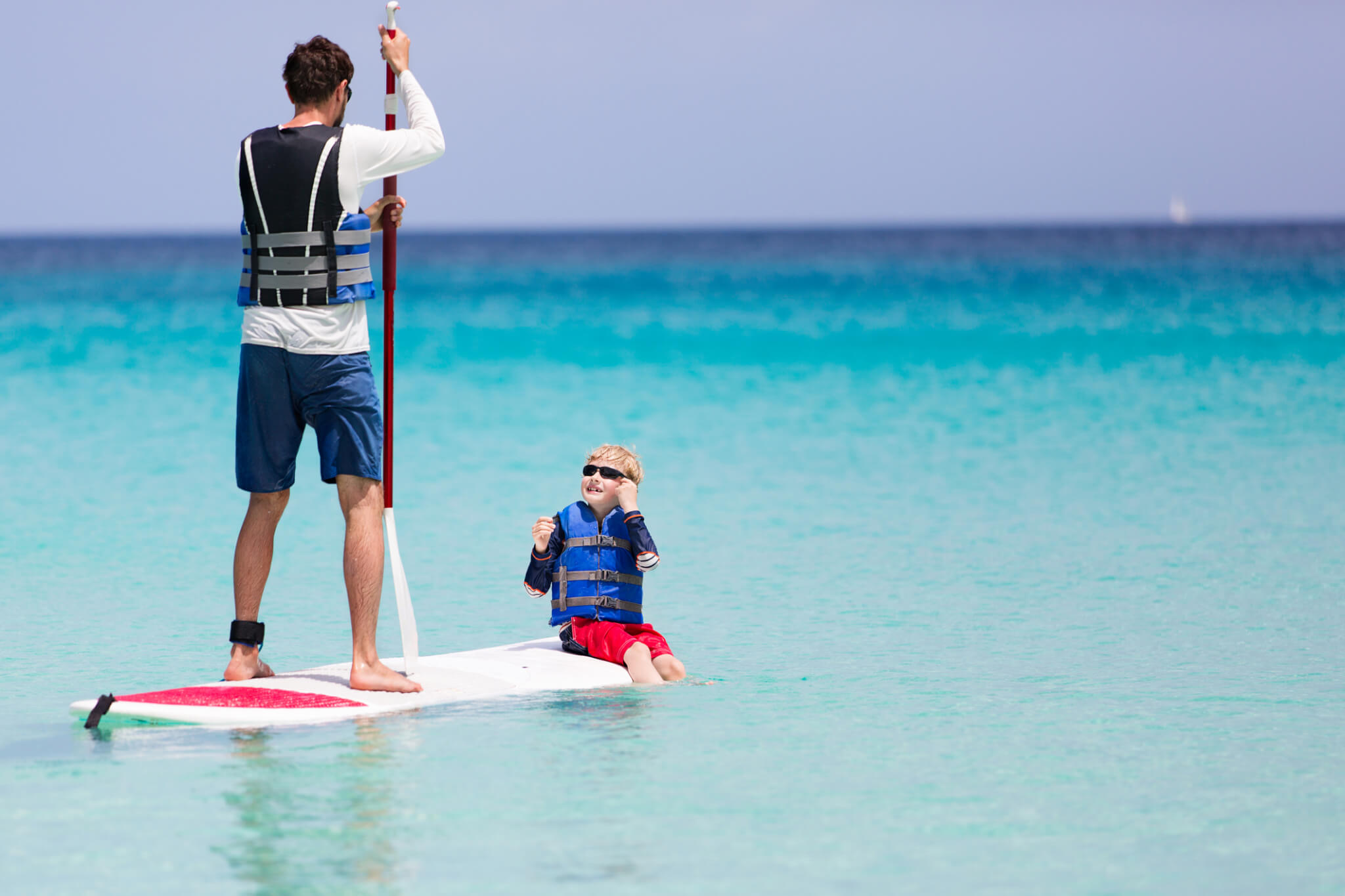 Man standing on a paddle board with a small child sat on the end, in clear blue water