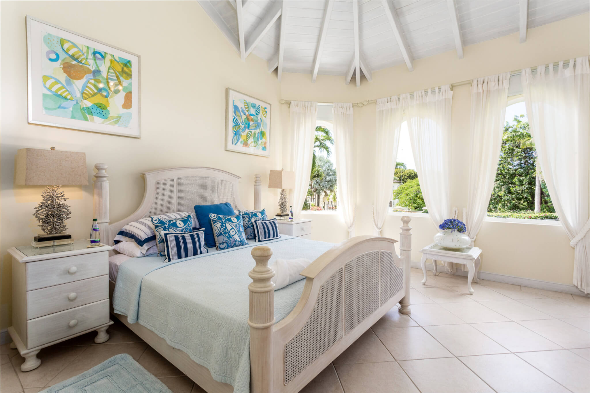 Bedroom with a large bed with light blue sheets and blue pillows. A high ceiling and multiple windows overlooking the garden